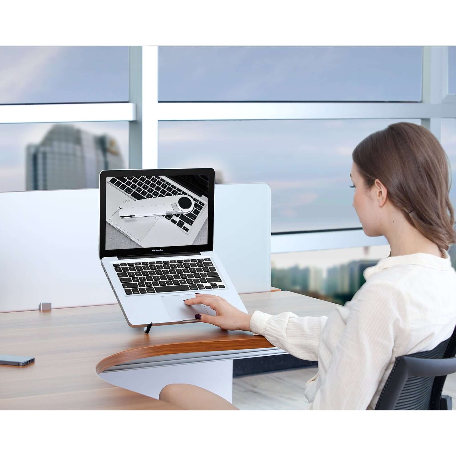 Woman using laptop on Spider laptop and tablet stand, white, at modern office desk with cityscape view in the background.