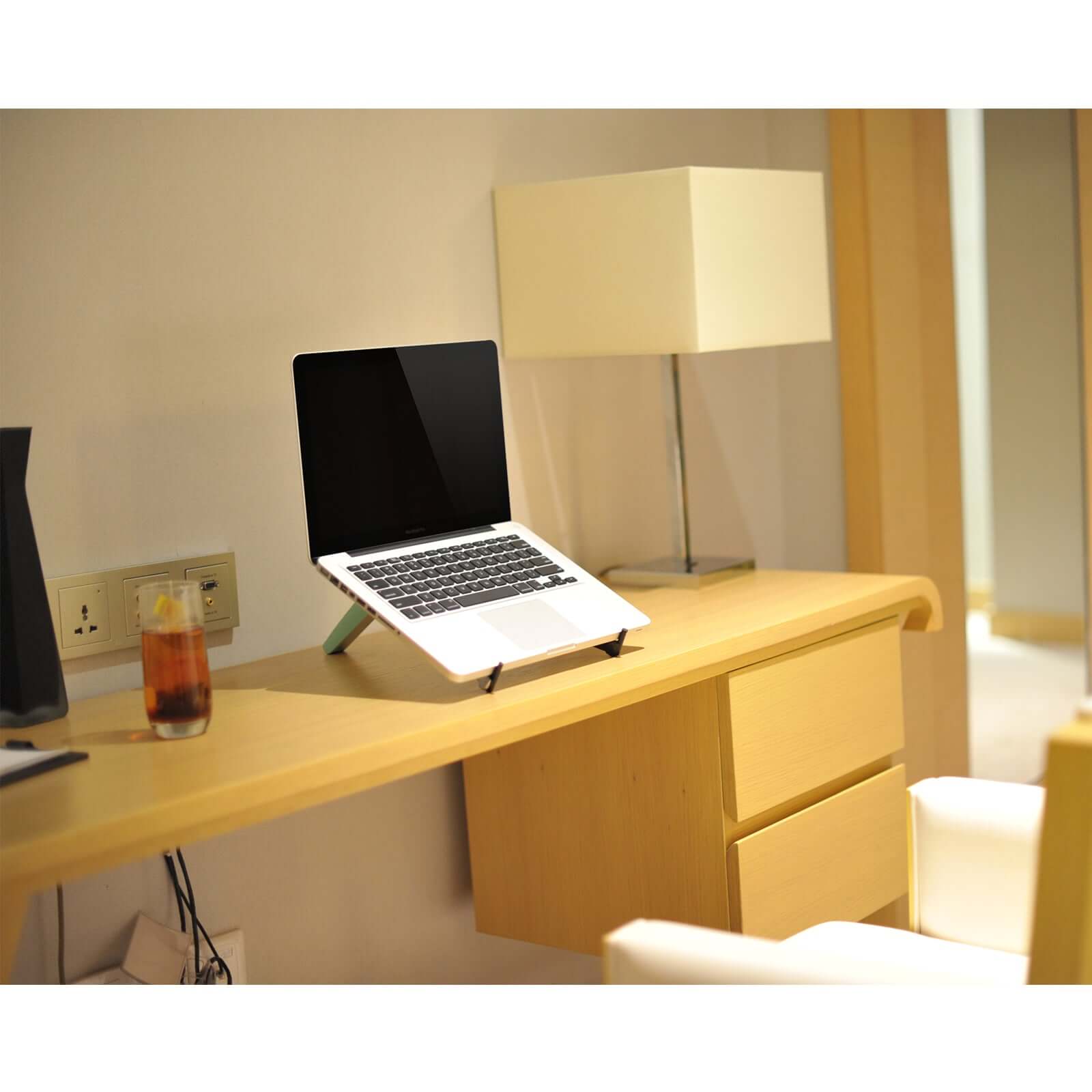 White spider laptop and tablet stand holding a laptop on a wooden desk with a lamp and a glass of beverage nearby.