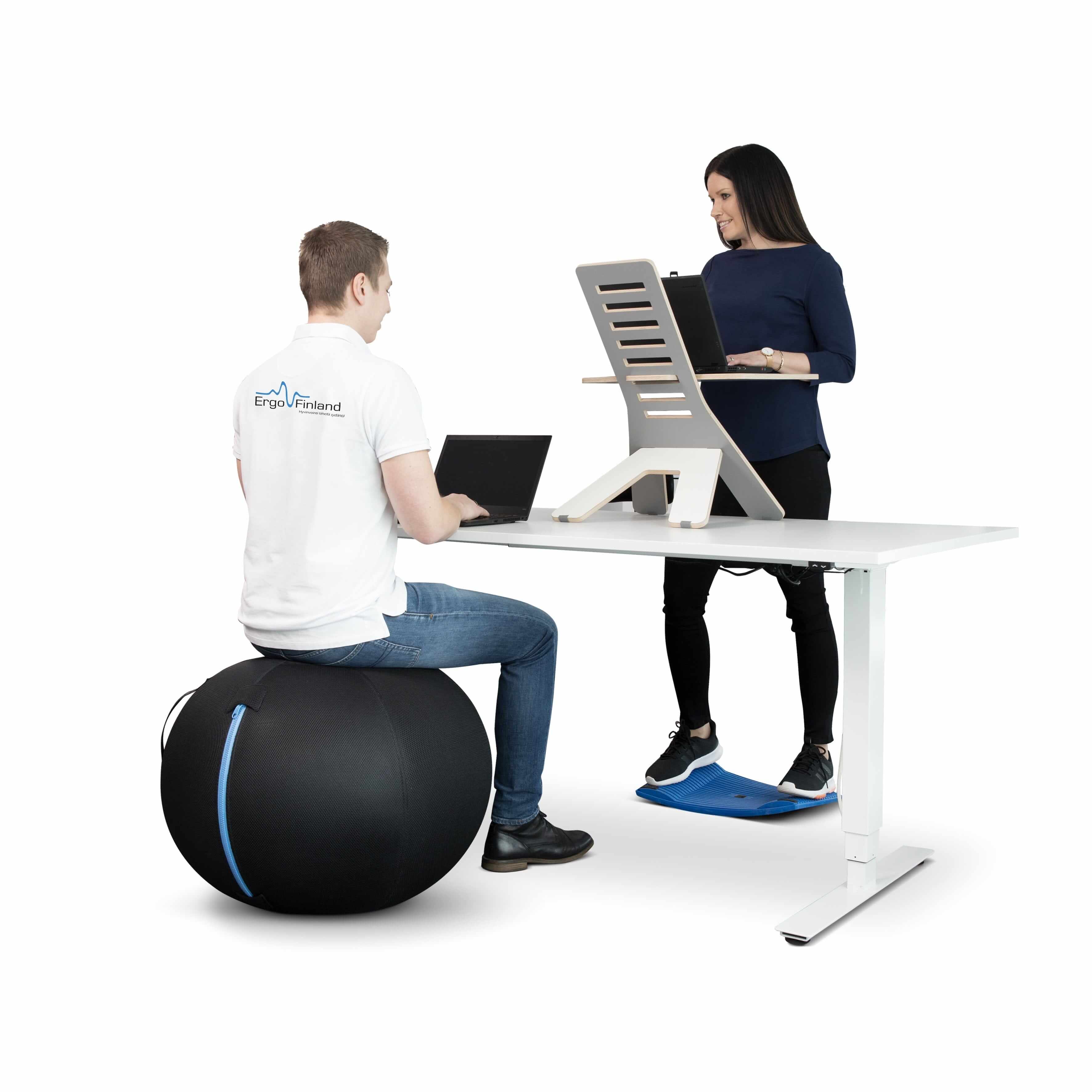 Person sitting on a GetUpBall office ball working on a laptop at a desk with a coworker standing next to them.